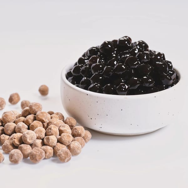 A white bowl filled with dark, glossy boba pearls next to a pile of light brown, uncooked boba.