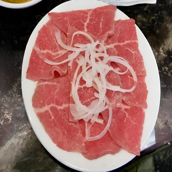 Thinly sliced raw beef with white onion slivers on a white plate, ready for Pho 07's Vietnamese hot pot.
