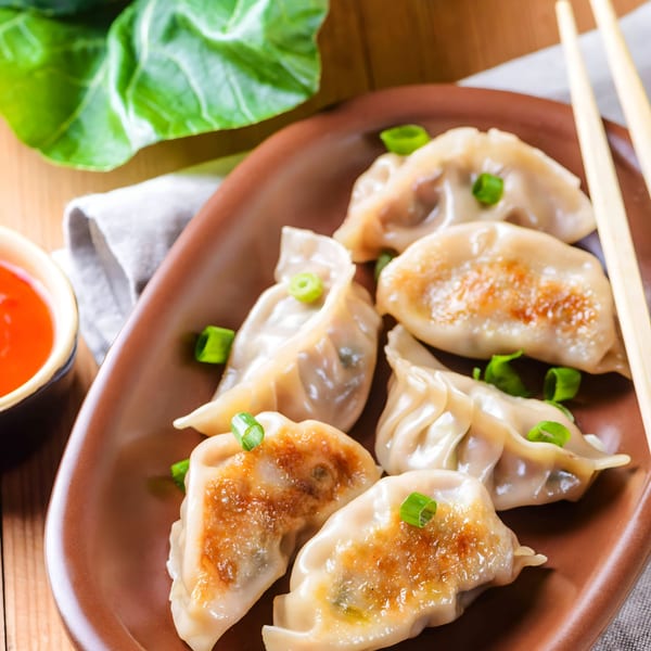 Pan-fried dumplings with green onions on a brown plate, with chopsticks and chili sauce.